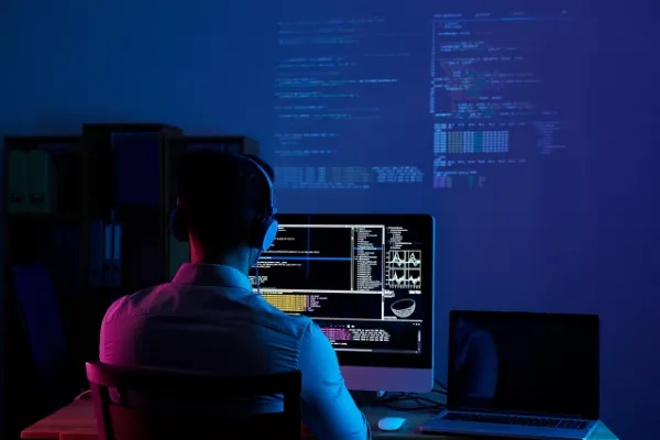 A person wearing headphones, seen from behind, working on a desktop computer with multiple screens displaying lines of code. The room is dimly lit with blue and purple ambient light, and code is projected onto the wall.