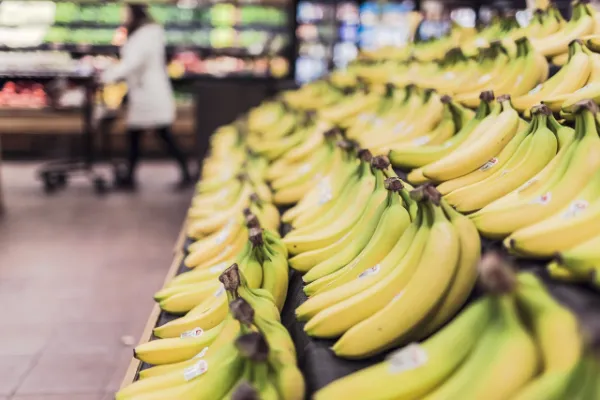 Rows of ripe yellow and slightly green bananas displayed in a supermarket produce section, with a blurred shopper and store interior in the background.