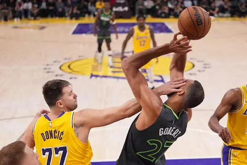 Luka Doncic of the Dallas Mavericks in a yellow jersey with number 77, drives to the basket while being defended by Rudy Gobert of the Minnesota Timberwolves in a green jersey with number 27, who is attempting to block the shot. Both players are looking at the basketball.