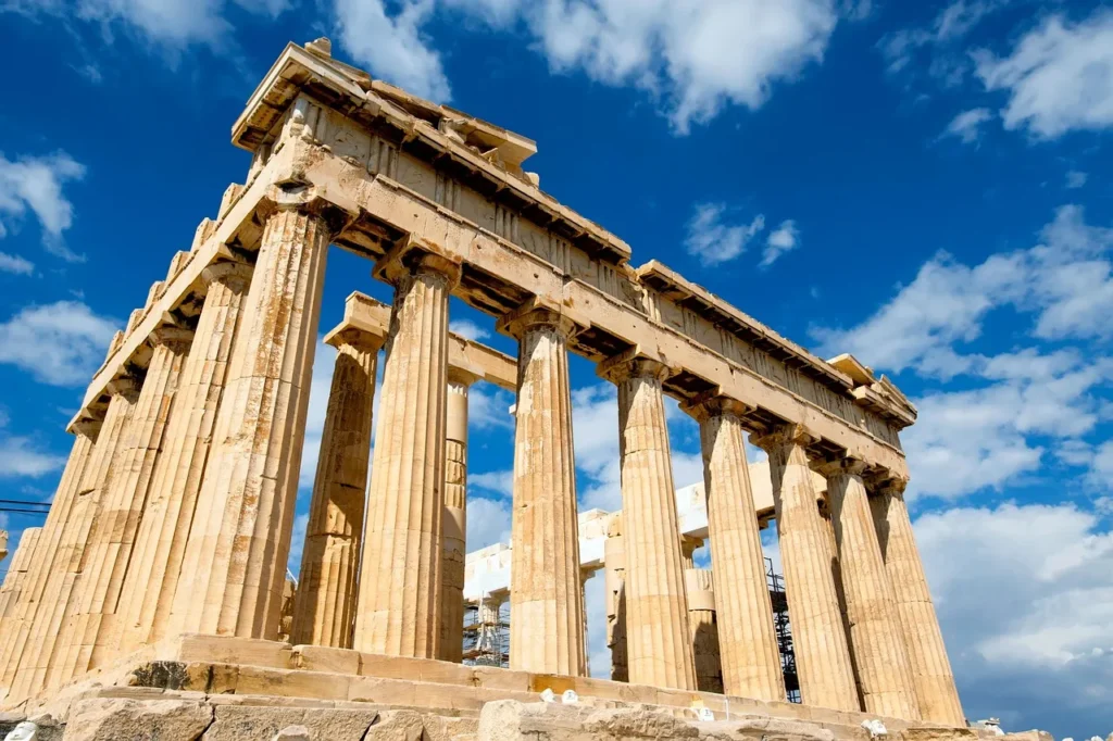 A low-angle view of the ancient Parthenon in Athens, Greece, showcasing its majestic Doric columns and classical architecture against a bright blue sky with scattered white clouds.
