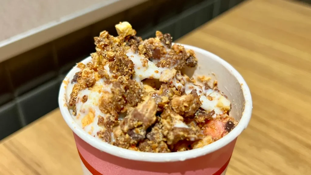 Close-up overhead shot of a McDonald's McFlurry in a white cup with a pink stripe, generously topped with crushed Kit Kat pieces and other mix-ins, showcasing a rich and textured dessert.