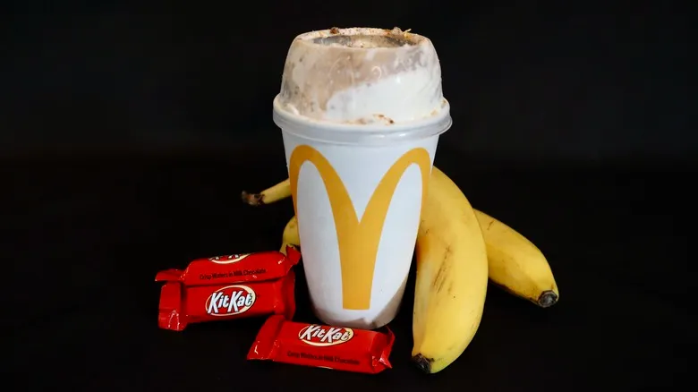 A McDonald's McFlurry cup filled with a creamy dessert, flanked by two ripe bananas and two individually wrapped Kit Kat chocolate bars, all against a dark background.