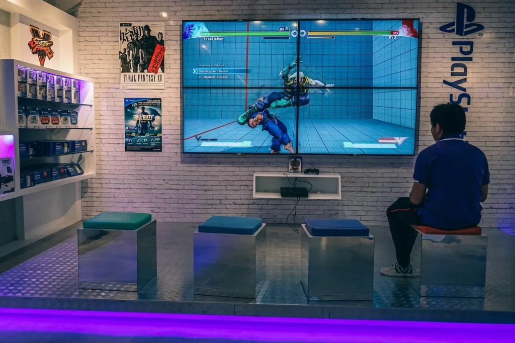 A person sits on a stool in a gaming setup, playing a fighting video game on a large screen, representing a casual gamer enjoying a potentially unblocked game.