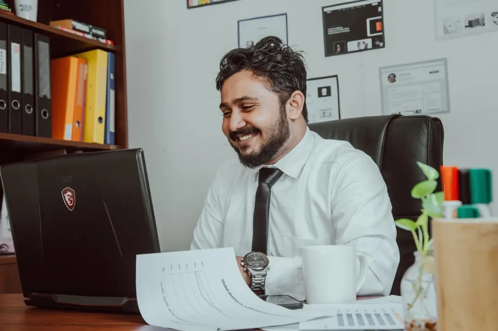 A smiling businessman with a beard, wearing a white shirt and black tie, seated at a wooden desk and looking happily at his laptop, with documents, a mug, and office decor in the background, suggesting a successful or pleasant work at highriskpay.com.