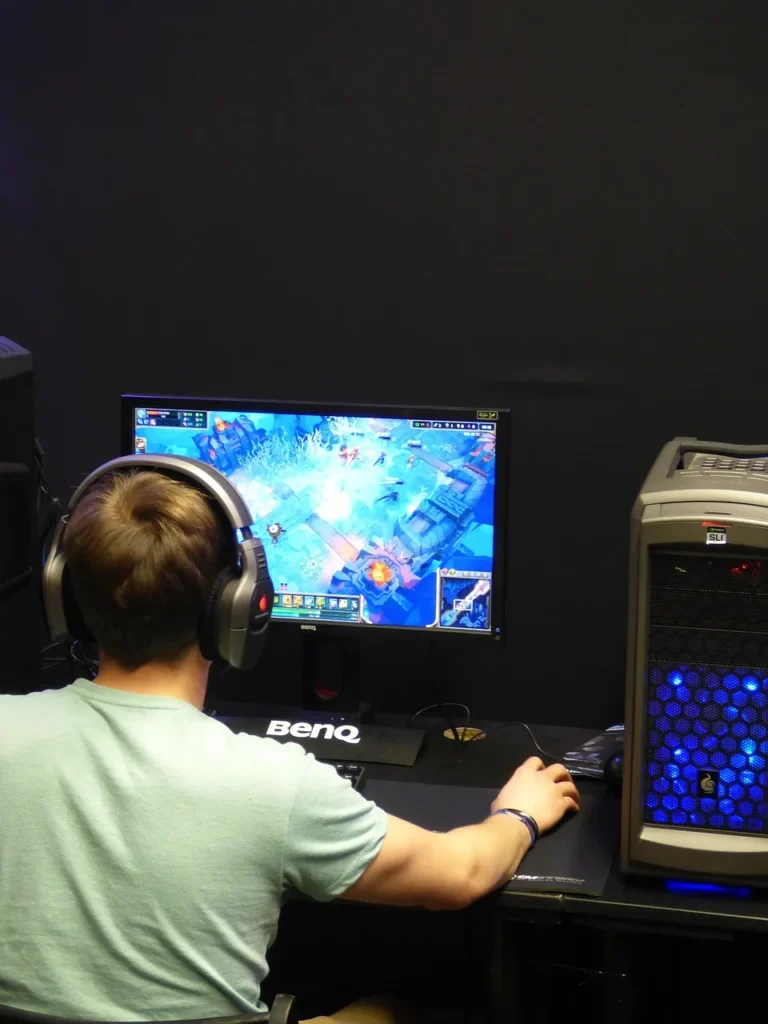 A gamer with large headphones on, playing a video game on a computer with a blue-lit tower.