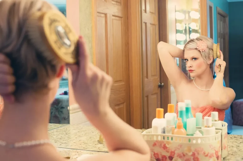 A woman in a peach dress and pearls brushes her short blonde hair while looking intensely at her reflection in a well-lit bathroom mirror.
