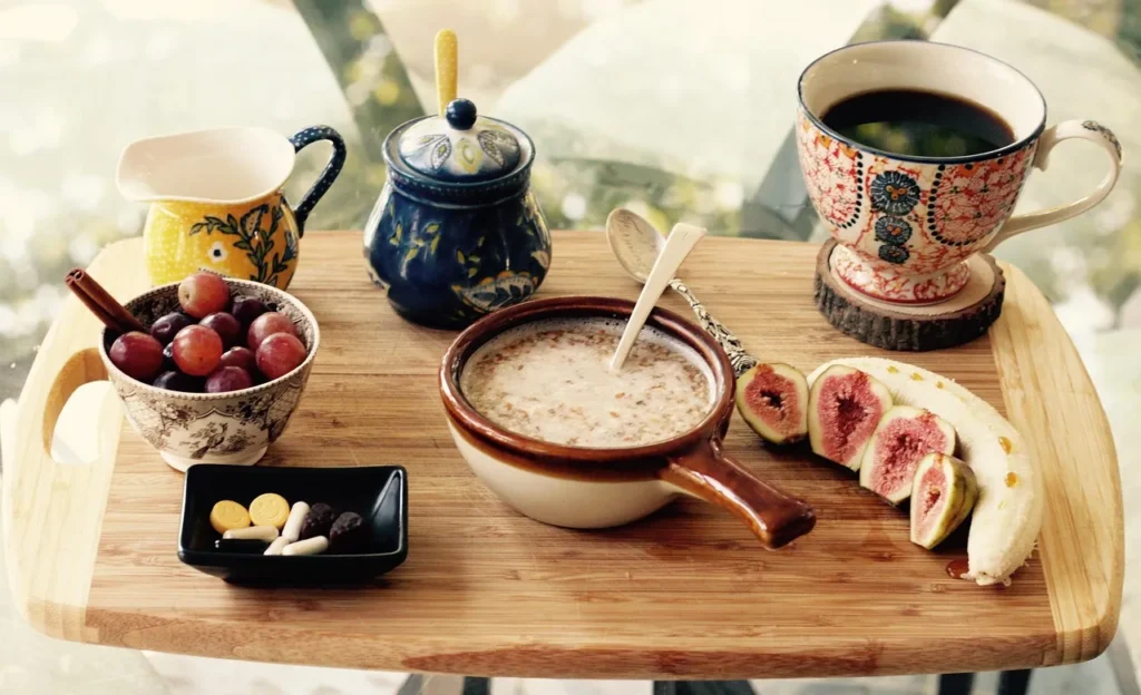 An appealing breakfast spread on a wooden cutting board featuring a bowl of cereal, sliced banana, halved figs, a bowl of grapes, and a cup of black coffee, symbolizing the common role of bananas in the daily diet.