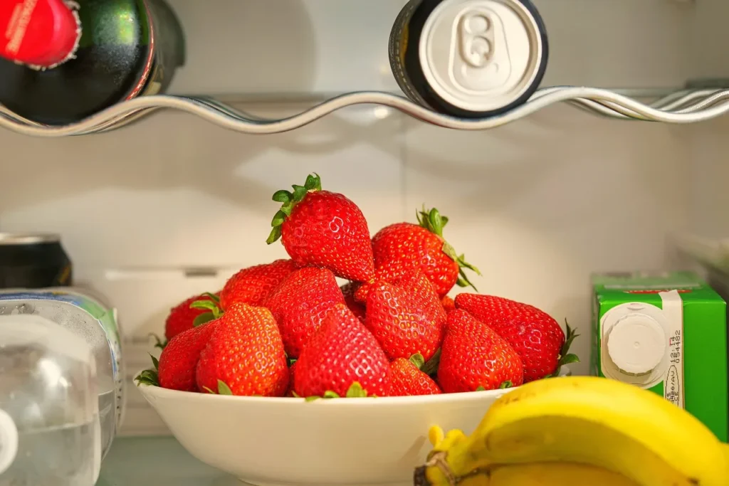 A close-up view inside a refrigerator showing a white bowl piled high with bright red strawberries, a cluster of yellow bananas in the foreground, and canned drinks on the shelf above.