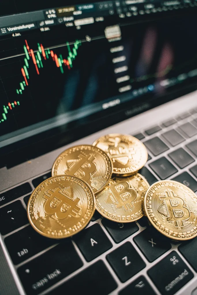Five physical gold Bitcoin coins stacked on a laptop keyboard with a green and red cryptocurrency price chart in the background.